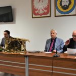 Trzech dorosłych mężczyzn w garniturach siedzących za prezydium na dużej sali. Na biurku znajduje się szopka.