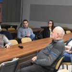 Grupa osób różnej płaci i wieku siedząca na rozległej sali konferencyjnej, Na biurku leży mikrofon.