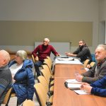 Grupa osób różnej płaci i wieku siedząca na rozległej sali konferencyjnej, Na biurku leżą dokumenty.