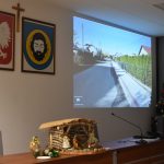 Ściana na którym eksponowana jest prezentacja z wyświetlacza. Obok widać krzyż i dwa herby.