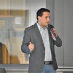 Dorosły mężczyzna w koszuli i garniturze przemawiający za pomocą miforonu na duzej sali.