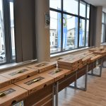 Paczki z kartonu lezące na podłużnym stole w okazałej sali konferencyjnej. W duże okna i widok na miasto,