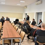 Grpa sosób różnej płci i wieku siedząca przy podłużnych stołach na sali konferencyjnej.