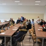 Duża grupa osób różnej płci i wieku siedząca na rozległej sali.