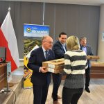 Trzech mężczyzn w garniturach wręczających kobiecie dwa kartonowe pudełka.