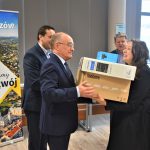 Trzech mężczyzn w garniturach wręczających kobiecie trzy kartonowe pudełka.