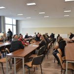 Duża grupa osób różnej płci i wieku siedząca na rozległej sali.