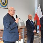 Trzech mężczyzn w garniturach stojących na sali. Jeden z nich przemawia za pomocą mikrofonu.