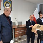 Trzech mężczyzn w garniturach wręcza kobiecie dwa kartonowe pudełka.