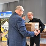 Wysoki, starszy mężczyzna wręcza młodszemu mężczyźnie w czarnej koszuli trzy kartonowe pudła.