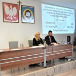 Kobieta i męzczyzna siedzących na prezydium sali konferencyjnej,. Na ścianie w tle widac zawieszone hreby oraz okno wyświetlacza.