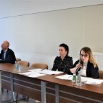 Dwie kobiety i męzczyzna ubrany w garnitur zasiadający przy podłużnym biurku na sali konferencyjnej. Na biurku leżą dokumenty oraz szklanki i butelki wody.