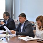 Trzech męzczyzn w garniturach oraz kobieta w białej koszuli siedzący przy biurkach na rozległej sali konferencyjnej.