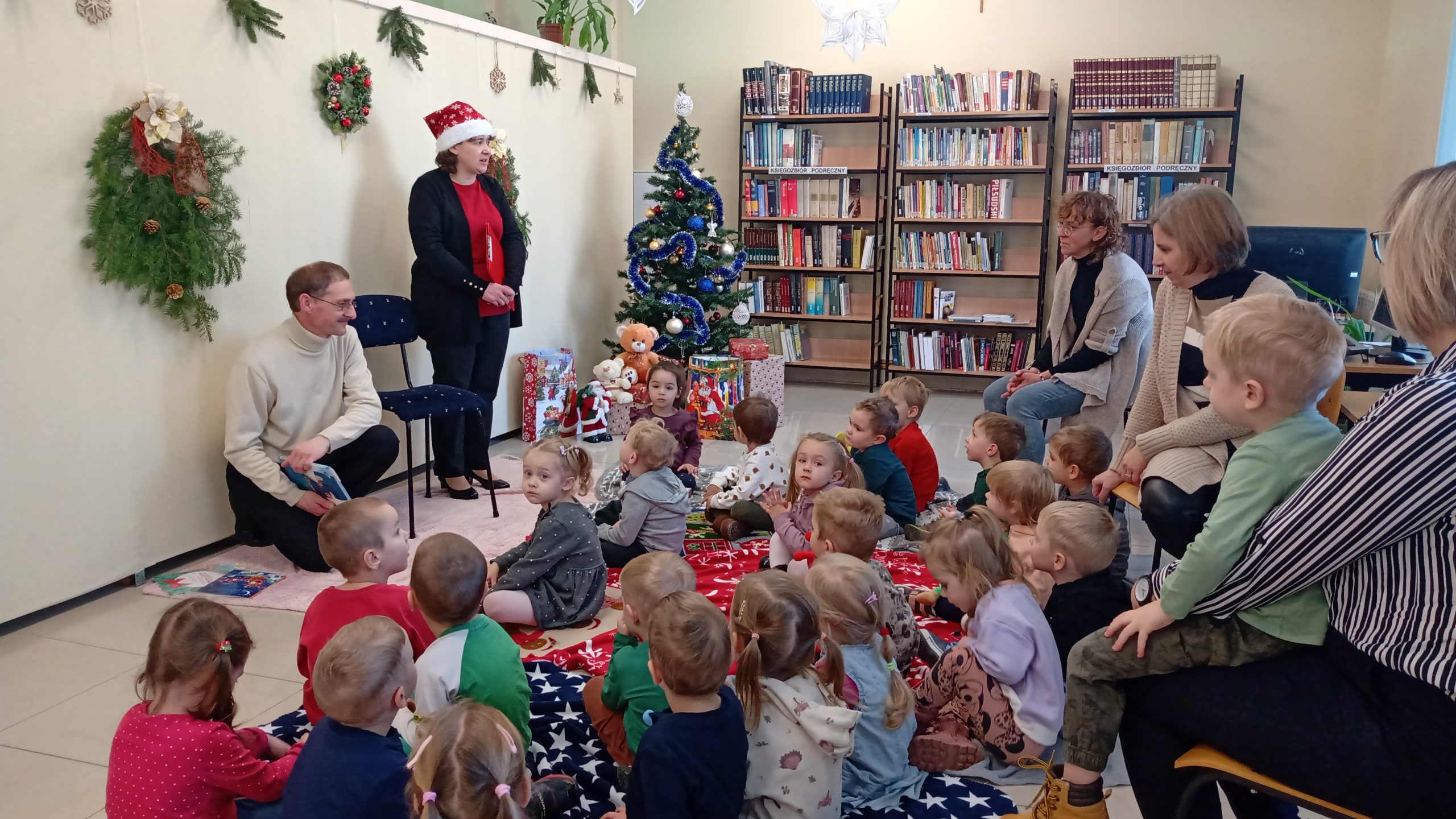 Grupa dzieci siedzi w bibliotece i słucha mężczyzny i kobiety opowiadającyh o prace w bibliotece.