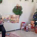 Grupa dzieci słucha mężczyzna, który siedzi na krzesle i opowiada im bajkę w bibliotece. Z boku stoi choinka ozdobiona.