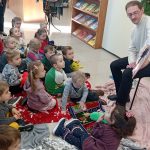 Grupa dzieci słucha mężczyzna, który siedzi na krzesle i opowiada im bajkę w bibliotece.