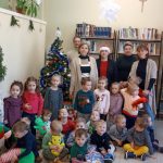 Grupa dzieci i kobiet pozują do zdjęcia w sali bibliotecznej.Za nimi półka z książkami.