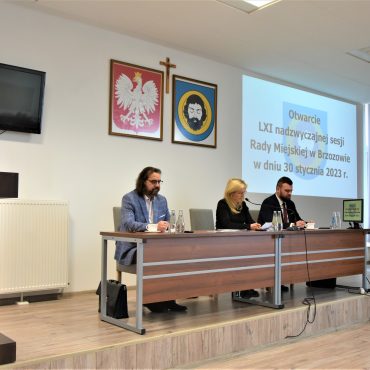 Kobieta i męzczyzna siedzących na prezydium sali konferencyjnej,. Na ścianie w tle widac zawieszone hreby oraz okno wyświetlacza.