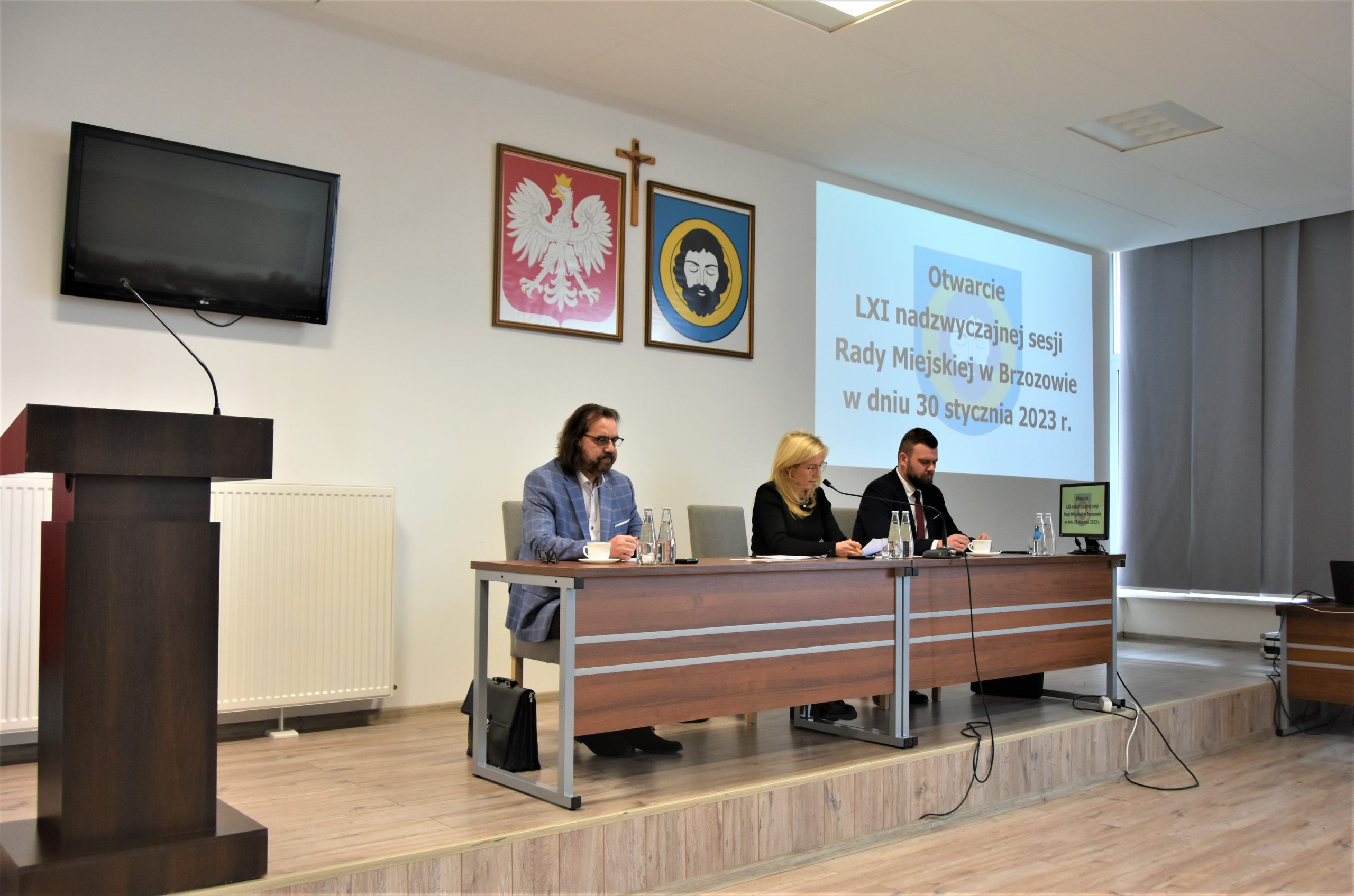 Kobieta i męzczyzna siedzących na prezydium sali konferencyjnej,. Na ścianie w tle widac zawieszone hreby oraz okno wyświetlacza.