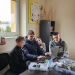 Grupa młodzieży z dorosłym mężczyzną spoglądają na laptop. Za nim leżą części elektroniczne.