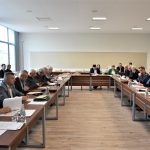 Grupa dorosłych osób w oficjalnych strojach siedzacych na sali obrad przy biurkach. Na blatach widać dokumenty, mikrofony, butelki wody ze szkalnkami.