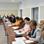 Grupa dorosłych osób w oficjalnych strojach siedzacych na sali obrad przy biurkach. Z tyłu widać poukładane drzwi i krzesła.