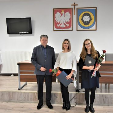 Mężczyzna w garniturze oraz dwie młode kobiety trzymające róże i ozdobne okładki. Pozujący do zdjęcia na sali gimnastycznej.