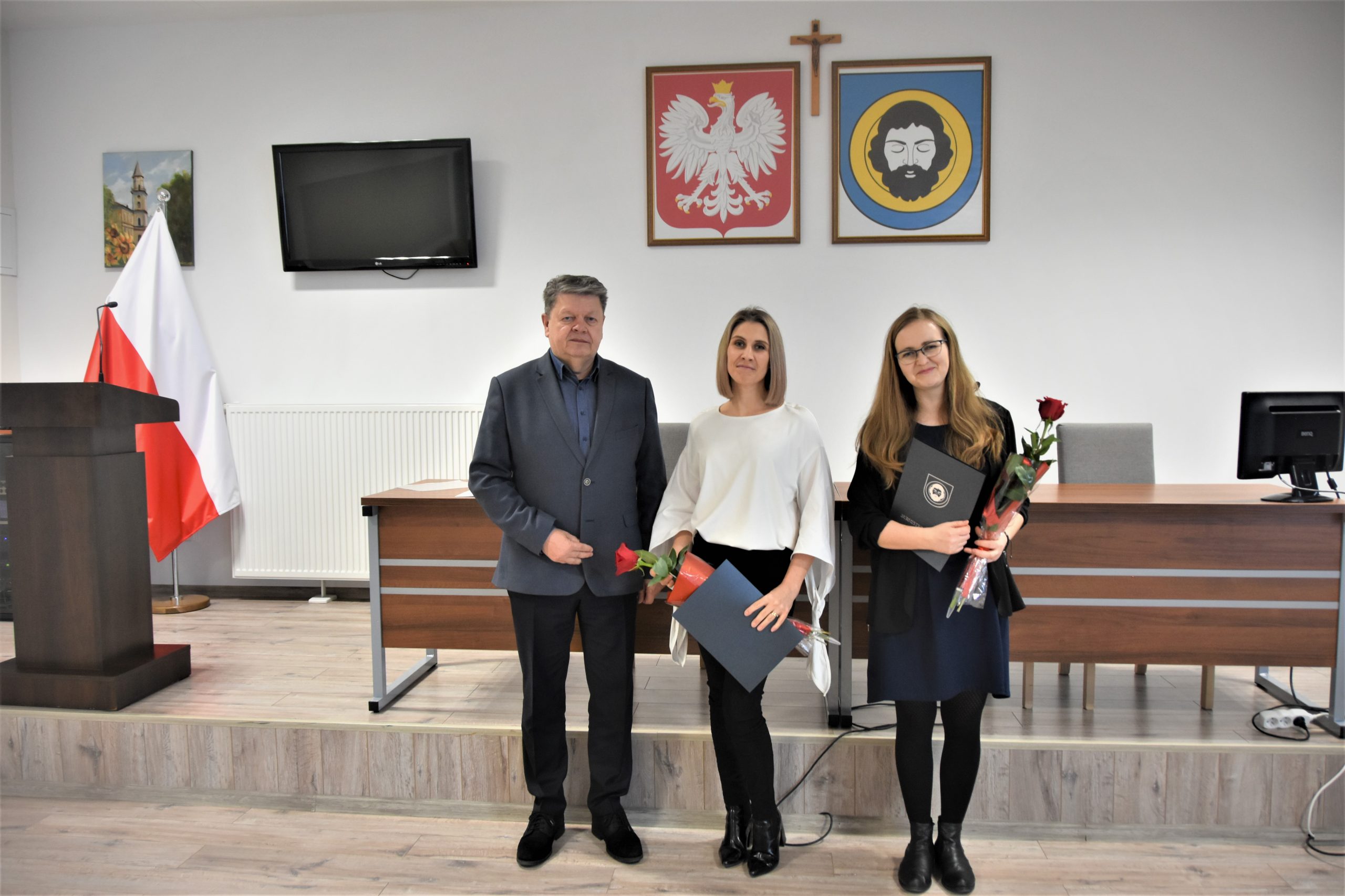 Mężczyzna w garniturze oraz dwie młode kobiety trzymające róże i ozdobne okładki. Pozujący do zdjęcia na sali gimnastycznej.