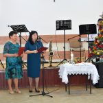 Dwie kobiety elegancko ubrane przemawiają do mikrofonu na sali. Obok nich stoi pięknie wystrojona choinka.