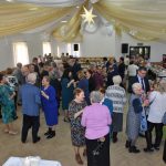 Grupa osób kobiet i mężczyzn dzieli się opłatkiem na sali.