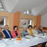 Grupa osób odświętnie ubranych siedzi przy stole nakrytym białym obrusem na sali.