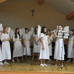 Grupa dziewczyn ubranych na biało ze skrzydełkami śpiewają na sali kolęde.