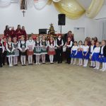 Grupa dzieci na sali śpiewa kolędę. Młody mężczyzna z boku trzymając w ręce torbę wręcza słodkości.