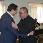 Młody mężczyzna w garniturze dzieli się opłatkiem z mężczyzną w sutannie.