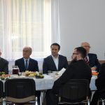 Grupa mężczyzn w graniturach siedzą przy stole nakrytym białym obrusem na sali.