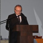 Mężczyzna w średnim wieku w graniturze stoi przy mównicy i mówi do mikrofonu.