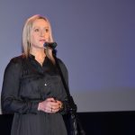 Kobieta w ciemnym czarnej sukni przemawia za pomoca mikrofonu do widowni.