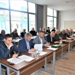 Grupa dorosłych osób w oficjalnych strojach siedzacych na sali obrad przy biurkach. Z tyłu widać okna z widokiem na miasto.