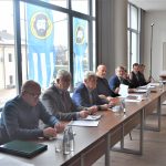 Grupa dorosłych osób w oficjalnych strojach siedzacych na sali obrad przy biurkach. Z tyłu widac okna z widokiem na miasto.