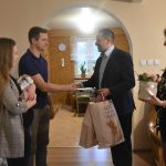 Młody mężczyzna w garniturze trzymający w rekach torby prezentowe gratluje młodemu meźczyźnie w koszulce. Obok nich stoi wysoka kobieta w marynarce o blond włosach trzymająca w rękach kosz z kwiatami, a druga kobieta w długich włosach trzyma na rękach dziecko.