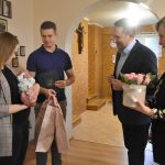 Młody mężczyzna w garniturze przygląda się młodym ludziom kobiecie i meżczyznie trzymających w rękach dziecko. Obok nich stoi kobieta trzymająca w rękach box z kwiatmi.