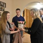 Kobieta w srednim wieku o blond włosach wręcza kobiecie o jasnych włosach box z kwiatami.