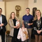 Cztery osoby stoją w jasnym pomieszczeniu. Mężczyzna w garniturze, kobieta o długich włosach trzymająca na rękach dziecko, młody mężczyzna w krótkiej koszulce, kobieta w marynarce o długich jasnych włosach.