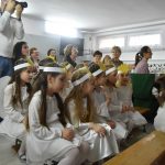 Grupa dzieci w stroju aniołków siedzących na ławkach w sali. Na około siedzą dorośli.