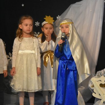 Grupa dzieci w strojach jasełkowych przy żłóbku. Jedna z dziewczynek w stroju Marii przemawia do mikrofonu.
