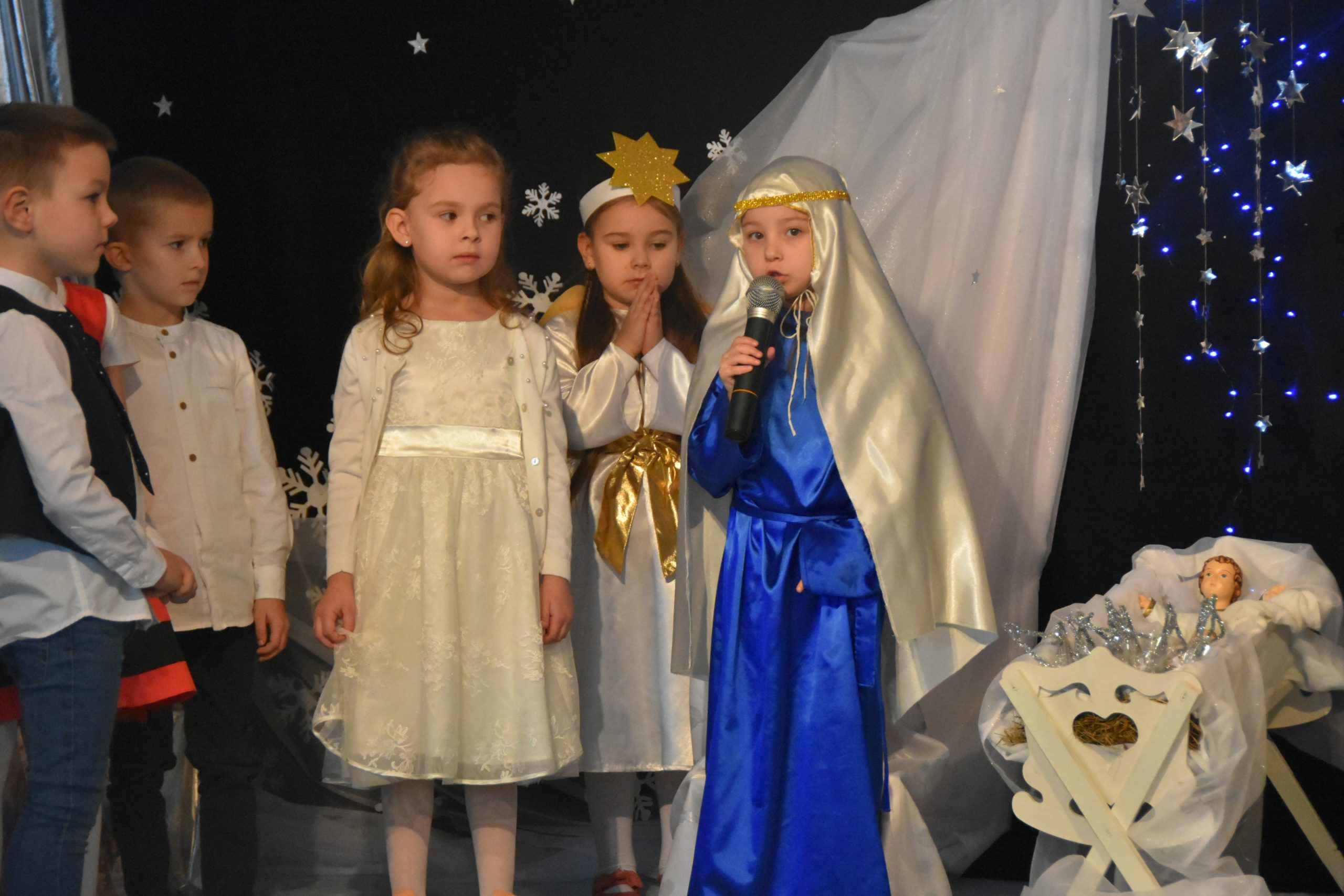 Grupa dzieci w strojach jasełkowych przy żłóbku. Jedna z dziewczynek w stroju Marii przemawia do mikrofonu.