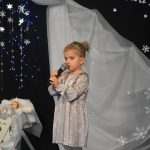 Dziewczynka w odświętnym stroju śpiewająca do mikrofonu przy żłóbku.