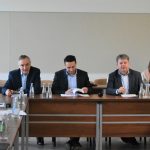 Trójka mężczyzn w garniturach i kobieta w białej bluzce zasiadający za długim biurkiem w sali konferencyjnej.