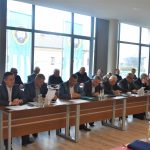 Grupa dorosłych osób różnej płci i wieku zasiadająca na dużej sali konferencyjnej.