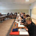 Grupa dorosłych osób różnej płci i wieku zasiadająca na dużej sali konferencyjnej. Na biurkach znajdują się dokumenty i woda.